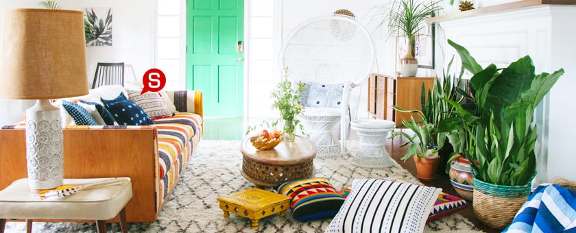 Un intérieur plein de couleurs dans le style boho avec un canapé confortable. Sur un tapis moelleux, il y a des coussins à motifs géométriques. Tout espace est rempli de belles décorations, de petites tables basses et quelques plantes vertes.