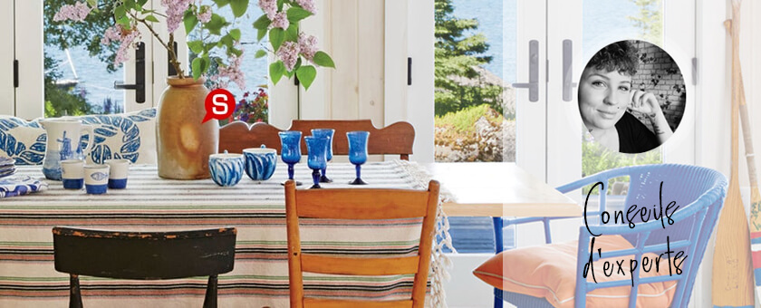 Une salle à manger bohème avec de grandes fenêtres. Sur le premier plan, il y une table à manger entourée des chaises en bois, chacune dans le style différent. Sur la table, il y a une nappe à rayures claire. Un vase de fleurs fraîches et quelques verres bleus.